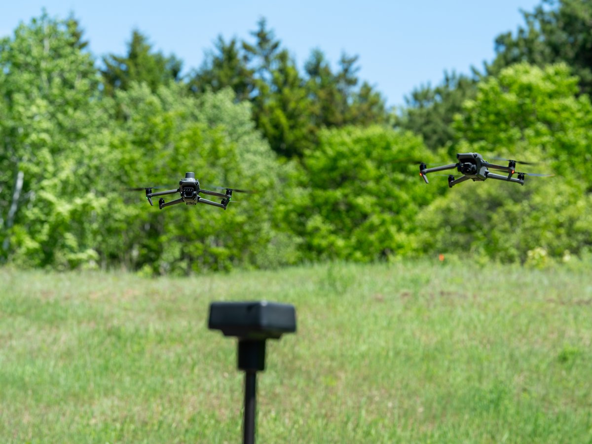 An RTK base station on a pole in a grassy field, with two drones flying behind it